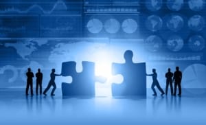 Silhouette of business people moving and joining pieces of jigsaw puzzle on a business and stock market blue background, with charts, diagrams, world maps and data arranged on grid and tables. World maps showing continents and countries with economy data and growth diagrams. Light glowing from the center. Copy space on bottom.