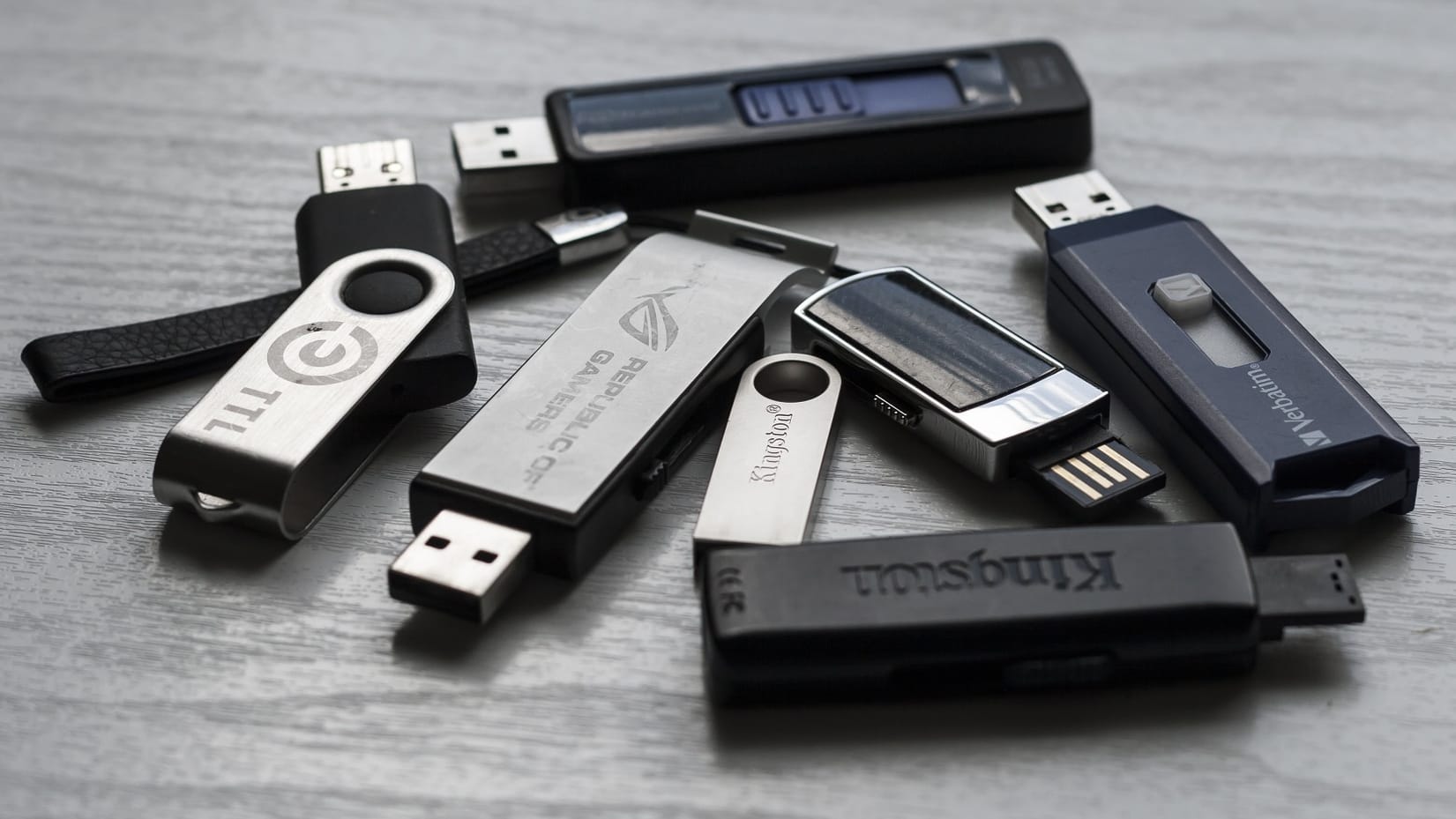 A scattered collection of various USB flash drives from brands like Kingston, Verbatim, and Republic of Gamers placed on a light wooden surface.