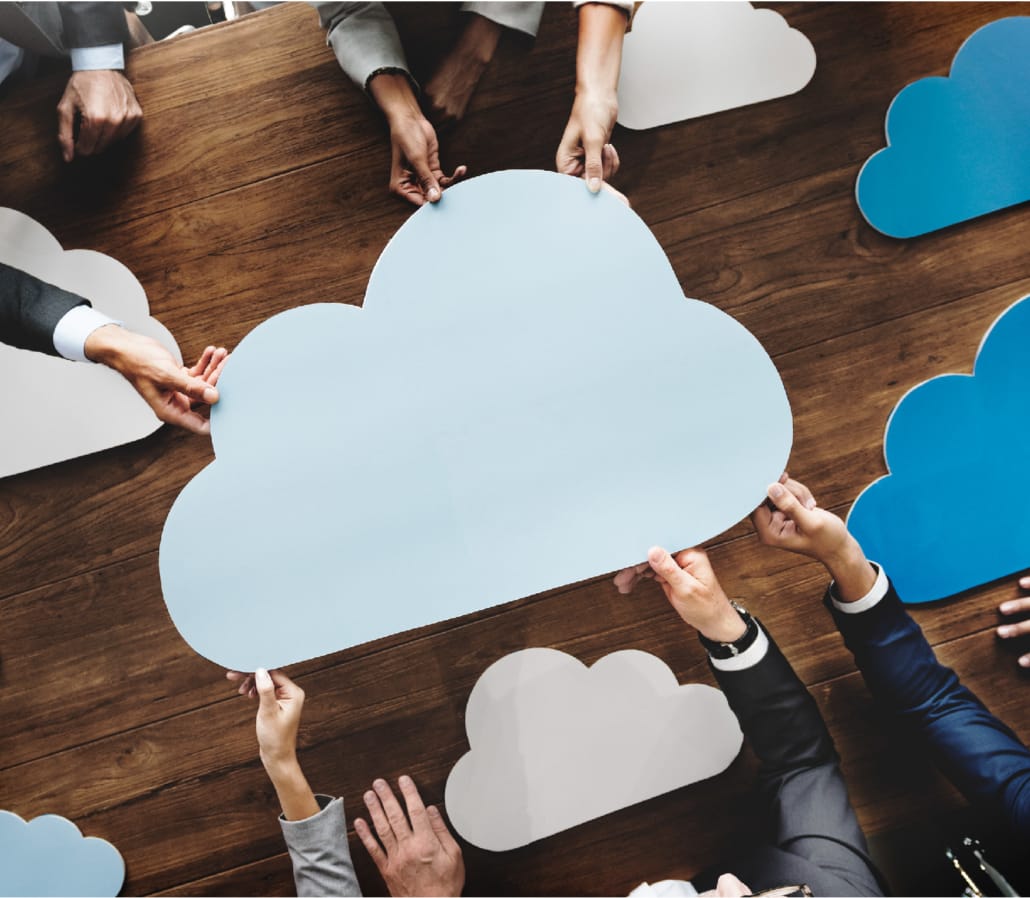 Group of people holding cutouts of cloud shapes over a wooden table, representing cloud computing collaboration.