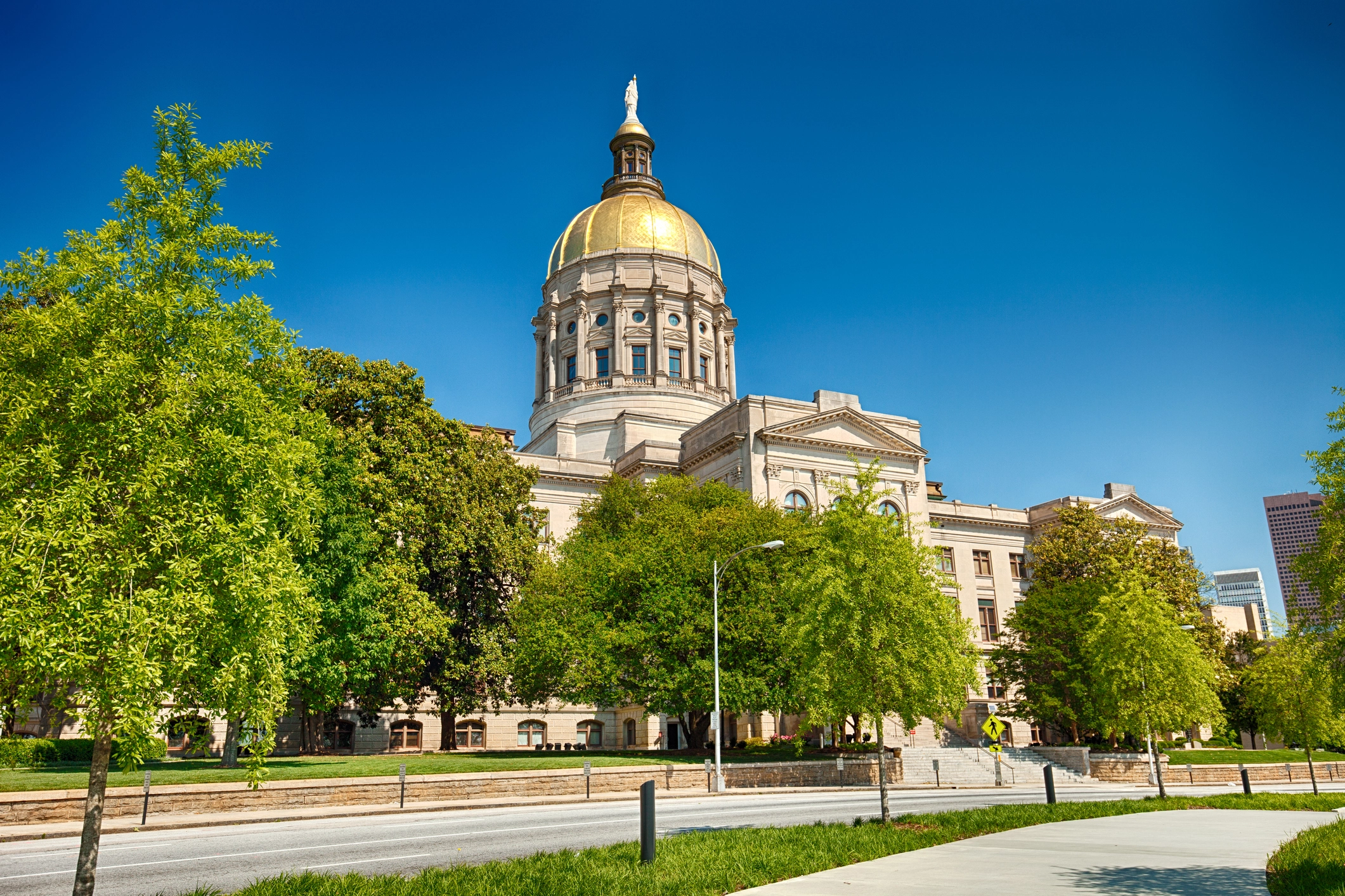 Exterior view of the Georgia State Capitol building with its iconic gold dome surrounded by lush green trees on a clear sunny day.