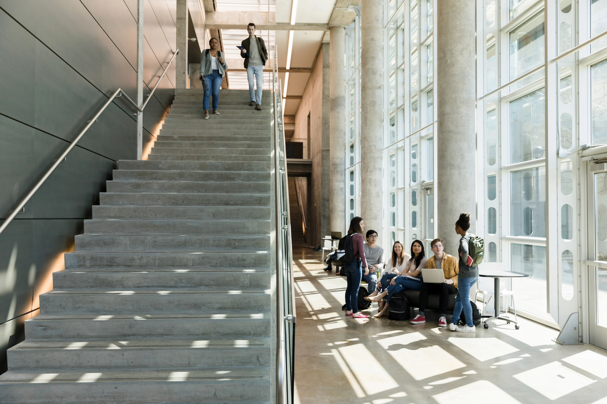 Group of college students socializing and studying near large windows in a modern campus building while two co-managed SOC students walk down a wide staircase.