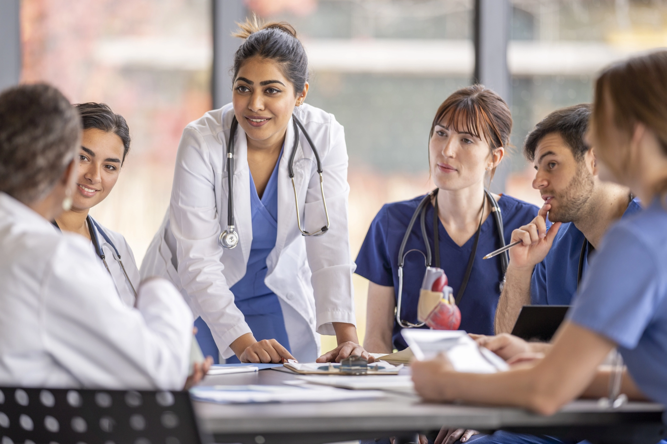 Diverse group of healthcare professionals in scrubs and lab coats gathered around a table in discussion during a medical team meeting.