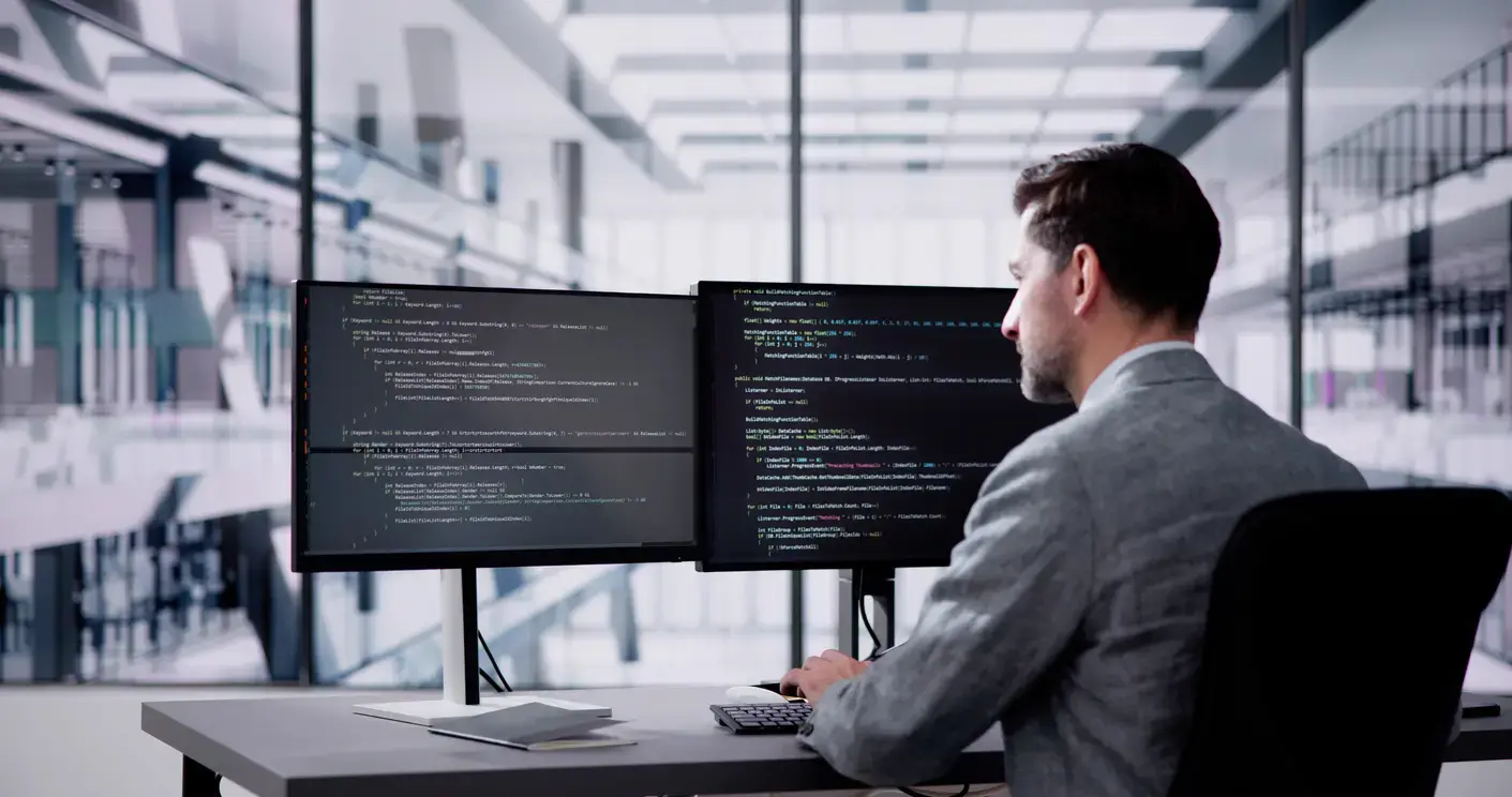 Enterprise IT security analyst reviewing code across dual monitors in a modern, well-lit office.
