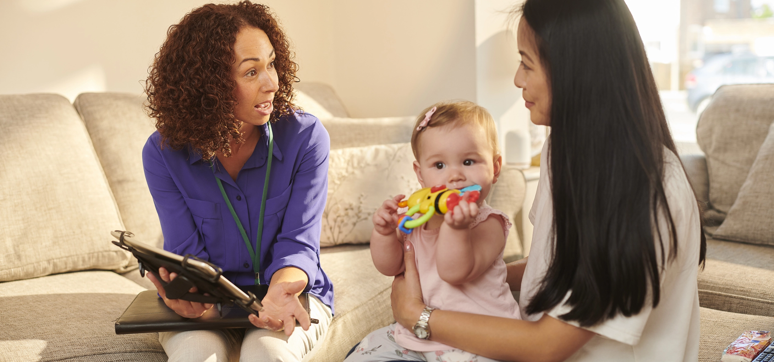 Female social worker with a tablet talking to a mother holding her baby on a couch during a home visit.