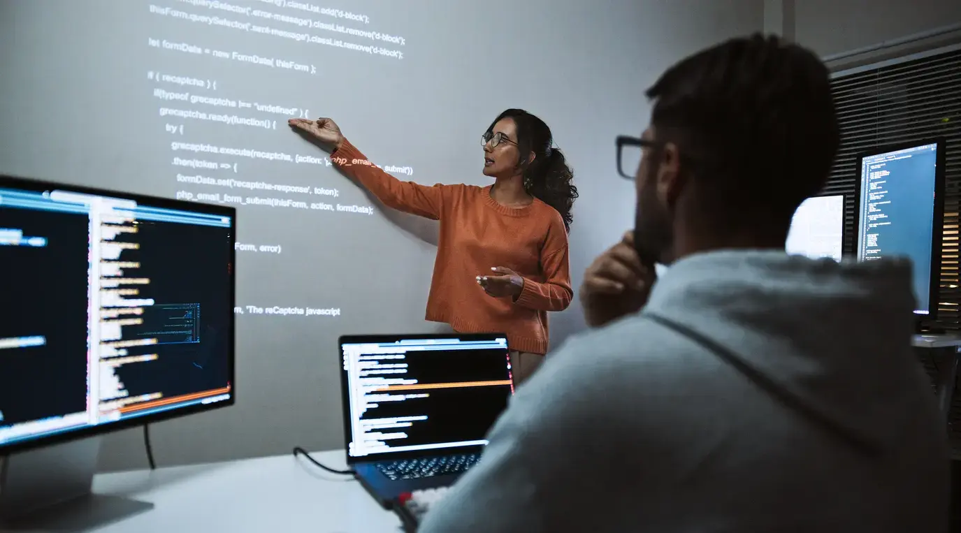 Woman presenting code on a wall-mounted screen to a colleague in a cybersecurity training session.