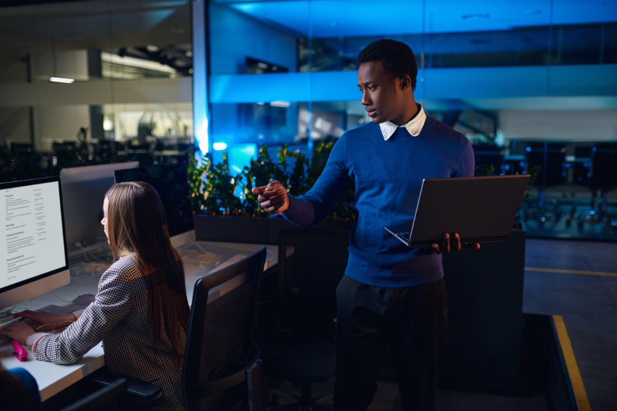 IT professional holding a laptop and monitoring activity on a workstation in a modern office, supporting a colleague.